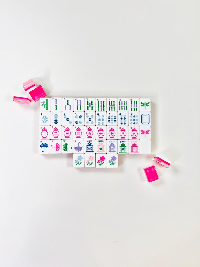May include: A set of white Mahjong tiles with colorful symbols and characters, including bamboo, dots, and dragons. The tiles are arranged on a white surface, with several pink and white tiles scattered around. The game pieces feature blue, green, pink, and purple accents.
