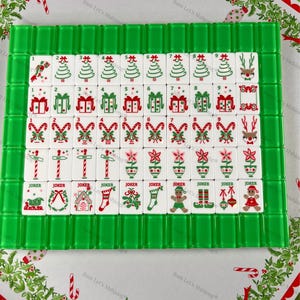Op de afbeelding: Een kerst-thema Mahjong-set met groene en witte tegels. De tegels tonen feestelijke afbeeldingen zoals kerstbomen, cadeaus en peperkoekenmannetjes. De set is gerangschikt op een decoratief oppervlak met vakantiepatronen.
