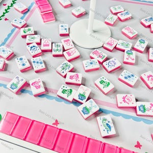 Puede incluir: Un juego de Mahjong completo con fichas rosas y blancas, con varios s&iacute;mbolos y dise&ntilde;os. El juego incluye un estante para las fichas y una superficie de juego. Las piezas del juego est&aacute;n dispersas sobre la superficie.