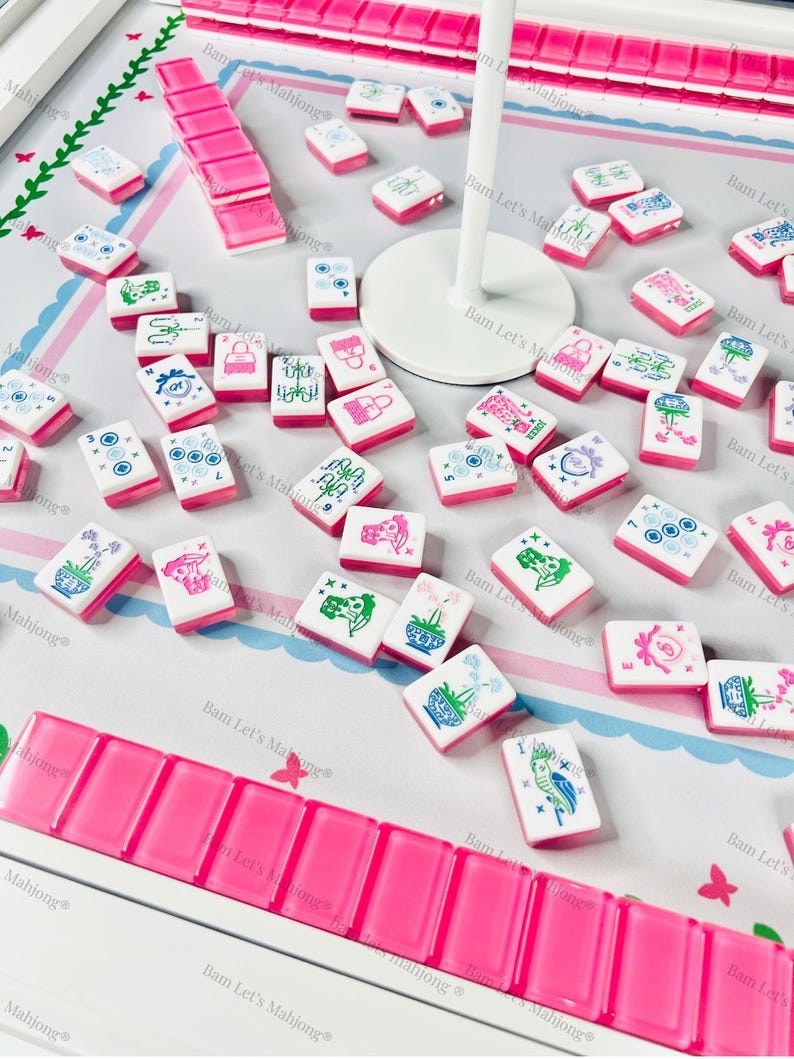 Puede incluir: Un juego de Mahjong completo con fichas rosas y blancas, con varios s&iacute;mbolos y dise&ntilde;os. El juego incluye un estante para las fichas y una superficie de juego. Las piezas del juego est&aacute;n dispersas sobre la superficie.