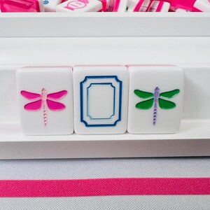 May include: White Mahjong tiles with pink and blue accents. Three tiles are in focus, each featuring a different design: a pink dragonfly, a blue frame, and a green dragonfly. Other tiles are visible in the background, all in a white tray.