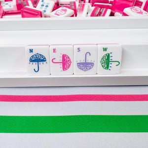May include: A collection of pink and white Mahjong tiles, with four tiles in the foreground displaying umbrella designs in blue, pink, purple, and green. The tiles are arranged on a white surface, with a striped fabric in the background.