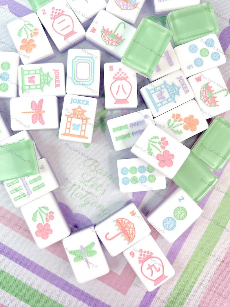 Op de afbeelding: Een verzameling witte Mahjong-tegels met diverse kleurrijke illustraties, waaronder bloemen, paraplu's en libellen. Sommige tegels hebben groene accenten, en sommige zeggen "Joker". De tegels liggen op een gestreepte achtergrond.