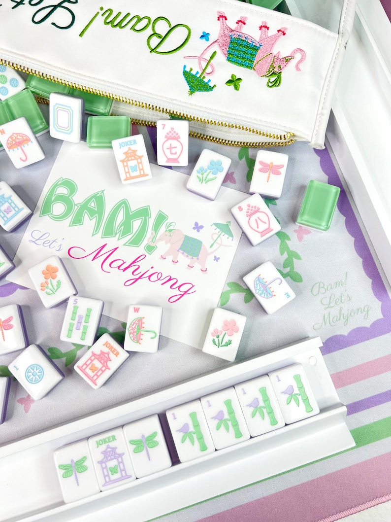 Op de afbeelding: Een Mahjong-set met witte tegels, elk versierd met verschillende symbolen en illustraties, waaronder bloemen, paraplu's en libellen. De set bevat een witte etui met ritssluiting met geborduurde details en de tekst "Bam! Let's Mahjong."