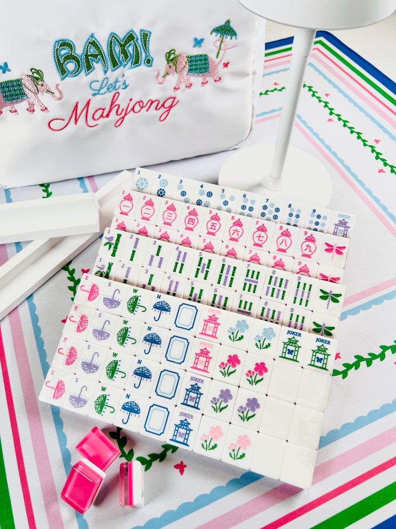 May include: A white Mahjong set with tiles featuring various symbols and colors, including pink, blue, and green. The set includes a white bag with the text "BAM! Let's Mahjong" and embroidered elephants. Also included are pink and white dice.
