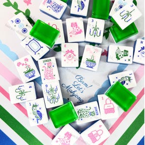 Pu&ograve; includere: Una collezione di tessere di Mahjong bianche con varie illustrazioni in rosa, blu e verde, tra cui fiori, animali e lampadari. Sono presenti anche pezzi rettangolari verdi. Il testo "Bam! Let's Mahjong" &egrave; visibile.