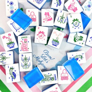 Peut inclure: Un ensemble de tuiles de Mahjong blanches avec diverses illustrations et des supports de tuiles en plastique bleu. Les tuiles pr&eacute;sentent des images d'animaux, de fleurs et de symboles en rose, vert et bleu. Le texte "Bam! Let's Mahjong" est visible.