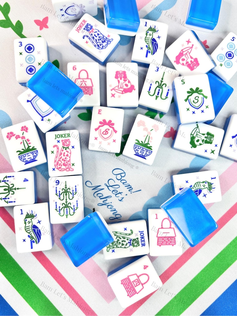 Peut inclure: Un ensemble de tuiles de Mahjong blanches avec diverses illustrations et des supports de tuiles en plastique bleu. Les tuiles pr&eacute;sentent des images d'animaux, de fleurs et de symboles en rose, vert et bleu. Le texte "Bam! Let's Mahjong" est visible.