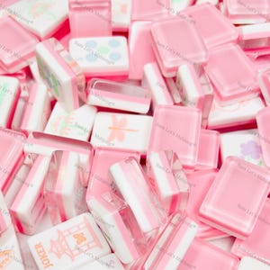 Puede incluir: Un conjunto de fichas de Mahjong rosas y blancas. Algunas fichas son de color rosa liso, mientras que otras tienen rayas blancas y rosas o im&aacute;genes decorativas. El texto "JOKER" es visible en una ficha. La imagen tiene el texto "Bam Let's Mahjong".