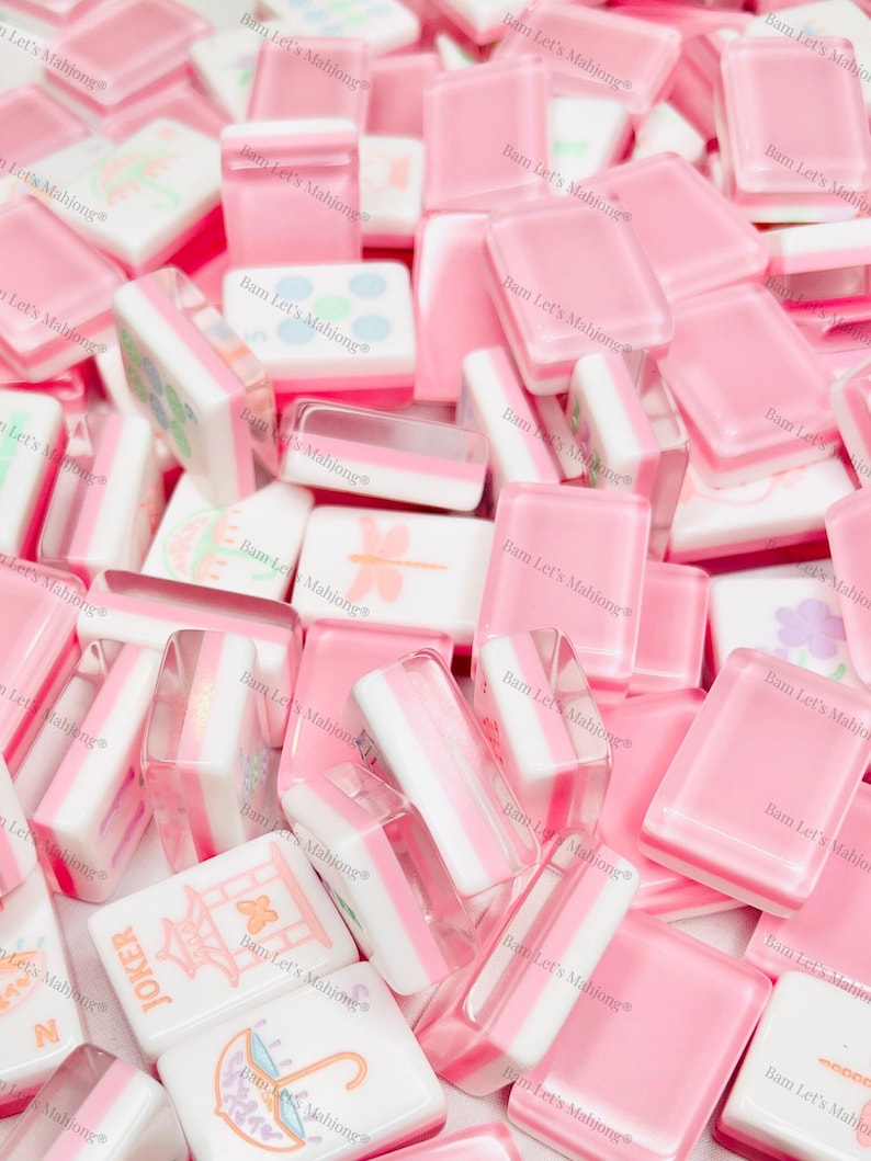 Puede incluir: Un conjunto de fichas de Mahjong rosas y blancas. Algunas fichas son de color rosa liso, mientras que otras tienen rayas blancas y rosas o im&aacute;genes decorativas. El texto "JOKER" es visible en una ficha. La imagen tiene el texto "Bam Let's Mahjong".