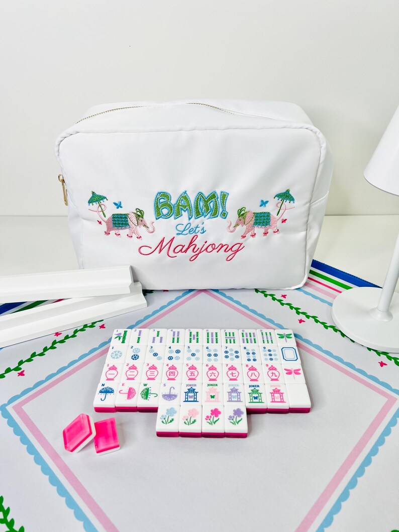 May include: A white Mahjong set with pink and white tiles, two pink dice, and a white zippered bag with embroidered elephants and the words "BAM! Let's Mahjong". The set is on a pink and white patterned surface.