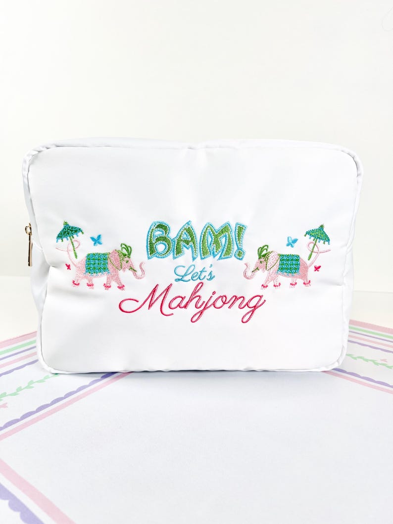 May include: White cosmetic bag with embroidered pink elephants, green umbrellas, and blue butterflies. The bag features the text "BAM! Let's Mahjong" in green and pink, with a gold zipper.