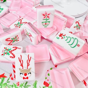 Op de afbeelding: Een verzameling roze en witte Mahjong-tegels met kerstthema's. Sommige tegels tonen zuurstokken, rendieren en bloempatronen. De tegels liggen op een wit oppervlak met groene en rode feestmotieven.