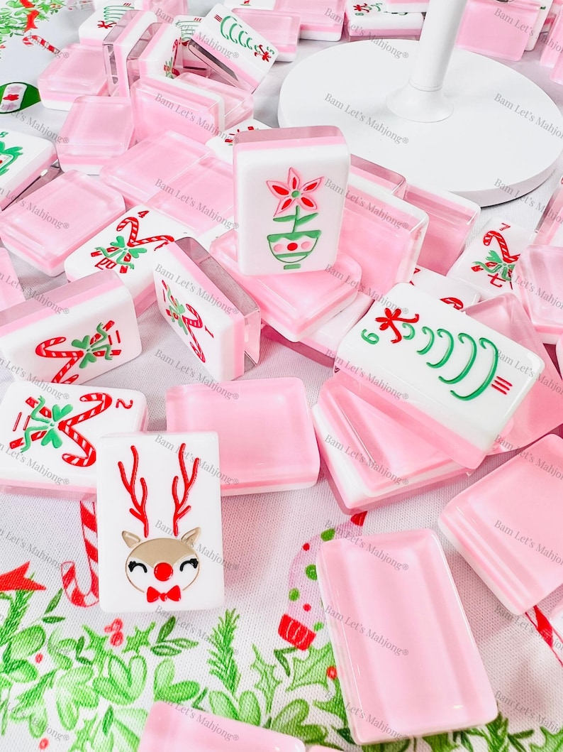 May include: A selection of pink and white Mahjong tiles, many with festive Christmas designs. The tiles display images of candy canes, reindeer, and flowers. The tiles are arranged on a white surface with seasonal green and red patterns.