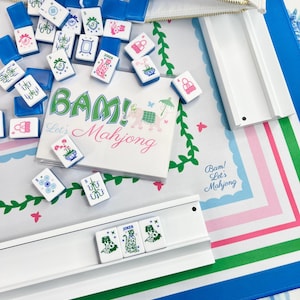 Peut inclure: Un &eacute;tui blanc zipp&eacute; d&eacute;verse des tuiles de Mahjong bleues et blanches sur un tapis &agrave; motifs. Les tuiles pr&eacute;sentent divers symboles et caract&egrave;res. Le tapis a un motif floral avec le texte "BAM! Let's Mahjong" en vert et rose.