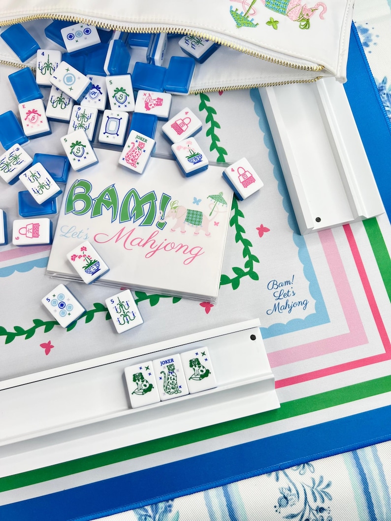 Peut inclure: Un &eacute;tui blanc zipp&eacute; d&eacute;verse des tuiles de Mahjong bleues et blanches sur un tapis &agrave; motifs. Les tuiles pr&eacute;sentent divers symboles et caract&egrave;res. Le tapis a un motif floral avec le texte "BAM! Let's Mahjong" en vert et rose.