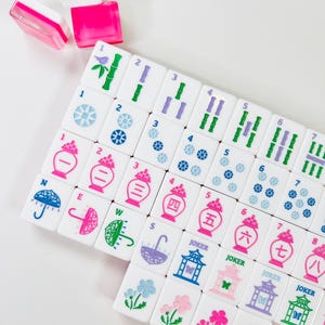 May include: A set of white Mahjong tiles with colorful illustrations and numbers. The tiles feature images of flowers, umbrellas, and bamboo. The set includes pink and clear plastic tile holders.