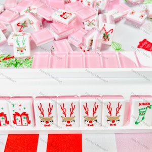 May include: A selection of Christmas-themed Mahjong tiles. The tiles display festive designs such as reindeer, gift boxes, and candy canes. The colour scheme includes white, pink, red, and gold accents, creating a cheerful holiday aesthetic.