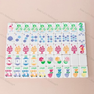 May include: A set of white Mahjong tiles with colorful illustrations. The tiles feature images of palm trees, seashells, coral, and seahorses. Some tiles have the word "JOKER" and images of golf carts and llamas.