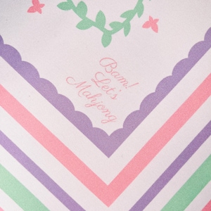 May include: A white textile with a pastel color scheme. The design features a chevron pattern in pink, purple, and green stripes. The top has a scalloped edge with text that reads "Bam! Let's Mahjong". Decorative elements include a green vine and pink butterflies.