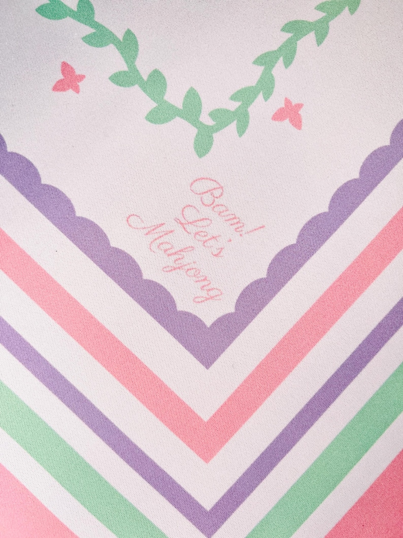 May include: A white textile with a pastel color scheme. The design features a chevron pattern in pink, purple, and green stripes. The top has a scalloped edge with text that reads "Bam! Let's Mahjong". Decorative elements include a green vine and pink butterflies.