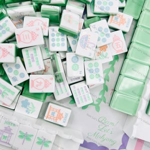 Op de afbeelding: Een verzameling witte en groene Mahjong-tegels met verschillende symbolen en cijfers. Sommige tegels hebben kleurrijke illustraties. De afbeelding bevat dobbelstenen en een zak met de tekst "Bam! Let's Mahjong".