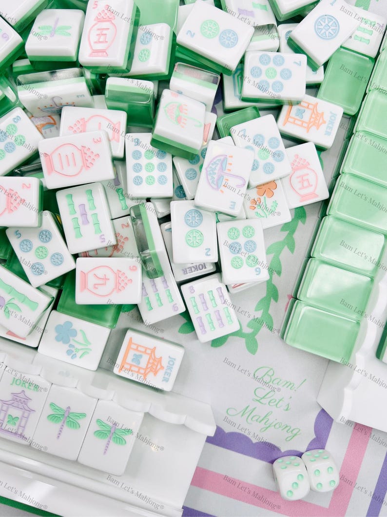 Op de afbeelding: Een verzameling witte en groene Mahjong-tegels met verschillende symbolen en cijfers. Sommige tegels hebben kleurrijke illustraties. De afbeelding bevat dobbelstenen en een zak met de tekst "Bam! Let's Mahjong".
