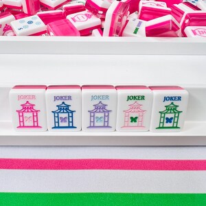 May include: A collection of pink and white Mahjong tiles, with a tray of tiles in the background. Five tiles in the foreground read "JOKER" and feature a pagoda design in pink, blue, purple, pink, and green. The background has a pink and green striped pattern.