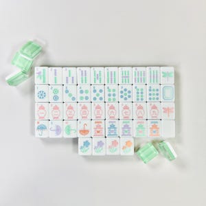 May include: A set of white Mahjong tiles with colorful symbols and numbers, including bamboo, circles, and characters. The tiles have pink, green, purple, and blue accents. Several translucent green and white tiles are scattered around the set.