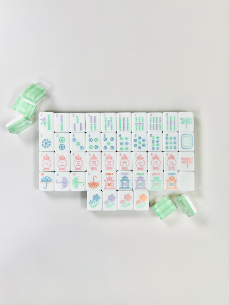 May include: A set of white Mahjong tiles with colorful symbols and numbers, including bamboo, circles, and characters. The tiles have pink, green, purple, and blue accents. Several translucent green and white tiles are scattered around the set.