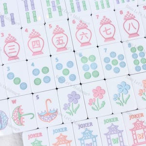 Puede incluir: Un conjunto de fichas de Mahjong blancas con varios s&iacute;mbolos y n&uacute;meros en verde, morado, rosa y azul. Las fichas presentan dise&ntilde;os de bamb&uacute;, flores y paraguas, junto con la palabra "JOKER".
