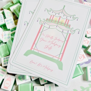 Op de afbeelding: Een verzameling Mahjong-tegels en twee kaarten. De kaarten tonen een pagode-ontwerp in groen en roze, met de tekst "I'm Silently Judging Your Mahjong Skills" en "Deal! Let's Mahjong". De tegels zijn wit en groen.