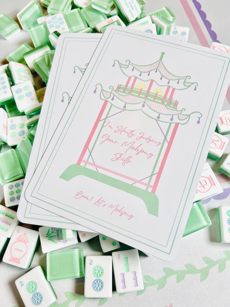 Op de afbeelding: Een verzameling Mahjong-tegels en twee kaarten. De kaarten tonen een pagode-ontwerp in groen en roze, met de tekst "I'm Silently Judging Your Mahjong Skills" en "Deal! Let's Mahjong". De tegels zijn wit en groen.