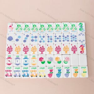 May include: A set of white Mahjong tiles with colorful illustrations. The tiles feature images of palm trees, seashells, coral, and seahorses. Some tiles have the word "JOKER" and images of golf carts and llamas.