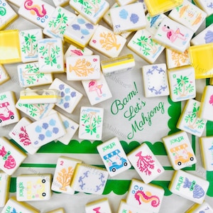 May include: A collection of white Mahjong tiles with colorful illustrations of coral, seahorses, and tropical scenes. Some tiles have the word "JOKER" on them. The tiles are scattered on a green and white patterned cloth.