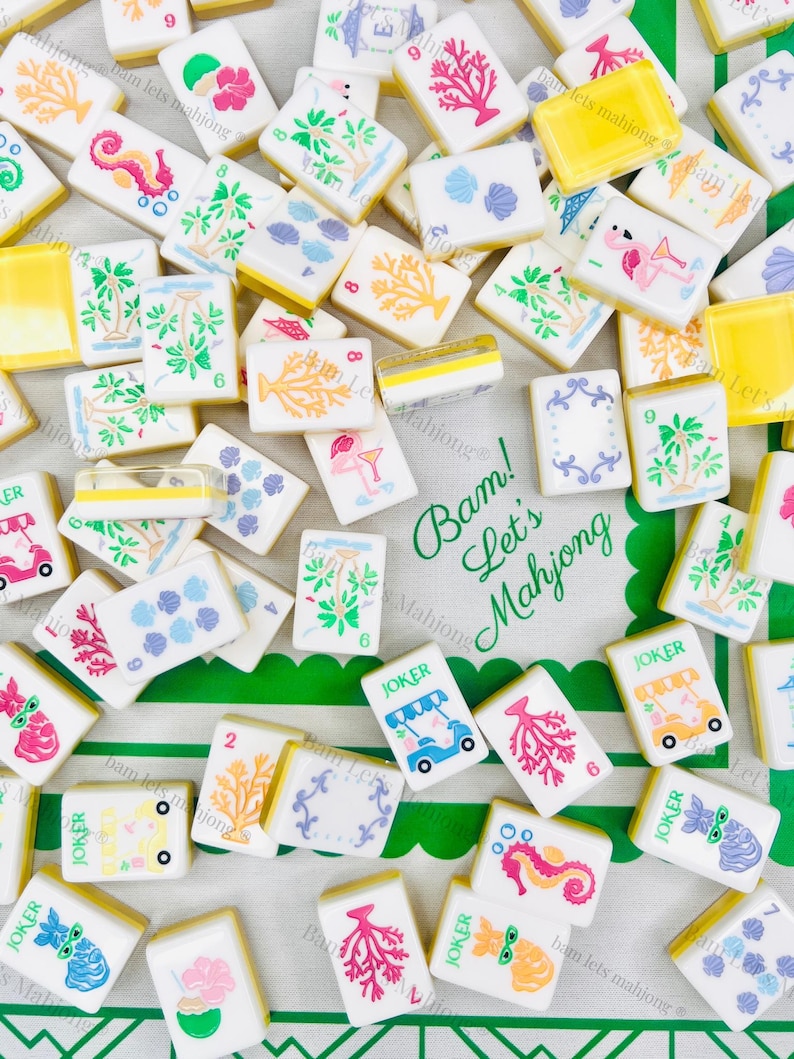 May include: A collection of white Mahjong tiles with colorful illustrations of coral, seahorses, and tropical scenes. Some tiles have the word "JOKER" on them. The tiles are scattered on a green and white patterned cloth.