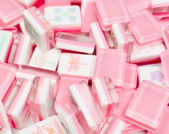 May include: A close-up shot of a pile of pink and white Mahjong tiles. Some tiles are solid pink, while others have white and pink stripes or small illustrations. The tiles are scattered, creating a visually appealing, textured pattern.