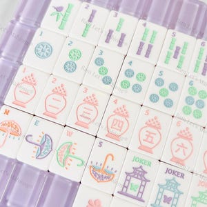 May include: A set of white Mahjong tiles with colorful illustrations and numbers. The tiles feature images of umbrellas, flowers, and pagodas, with a purple tile holder. The set includes tiles marked with the word "Joker".