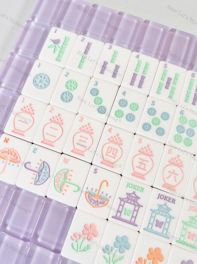 May include: A set of white Mahjong tiles with colorful illustrations and numbers. The tiles feature images of umbrellas, flowers, and pagodas, with a purple tile holder. The set includes tiles marked with the word "Joker".