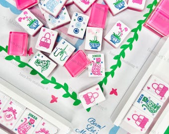 Puede incluir: Un conjunto de fichas de Mahjong y soportes rosas sobre una tela estampada. Las fichas presentan varios símbolos e ilustraciones en rosa, azul y verde. El texto "Bam! Let's Mahjong" es visible en la tela.