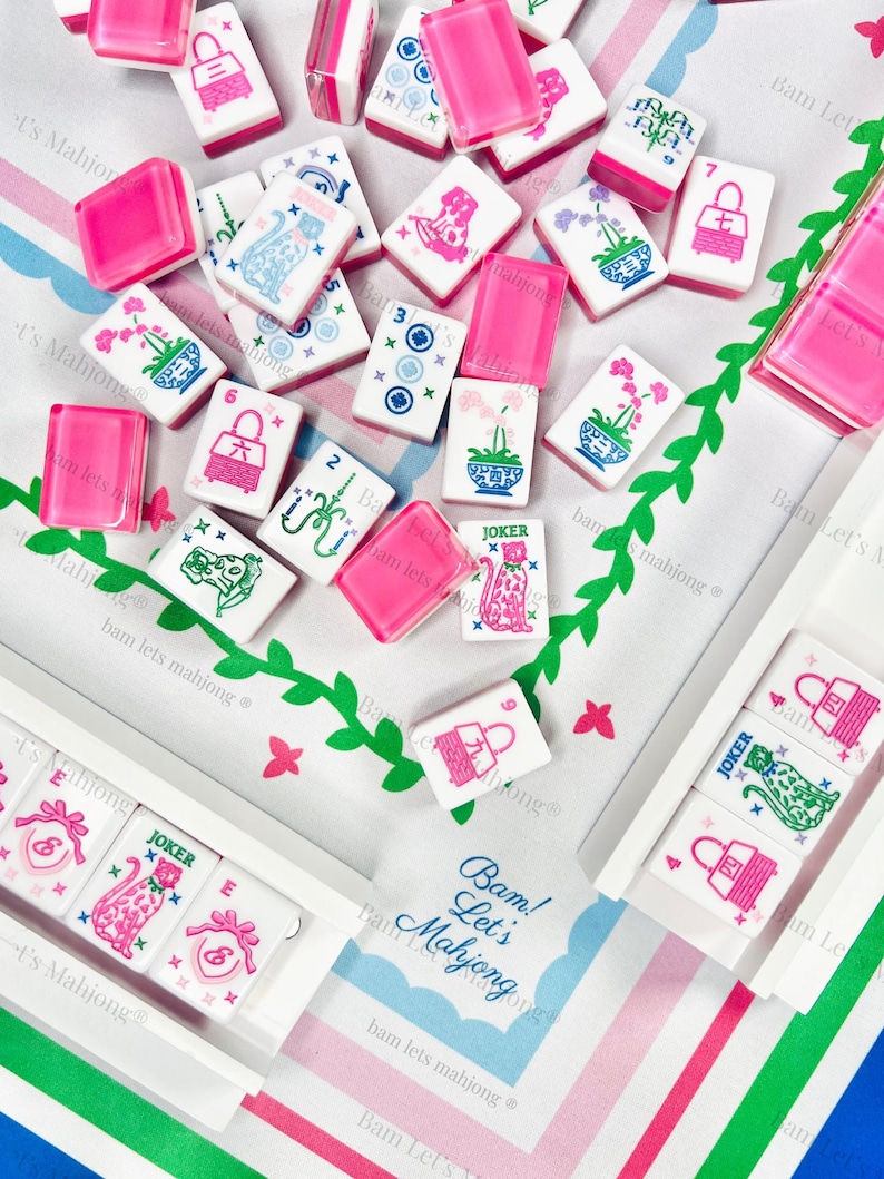 Puede incluir: Un conjunto de fichas de Mahjong y soportes rosas sobre una tela estampada. Las fichas presentan varios s&iacute;mbolos e ilustraciones en rosa, azul y verde. El texto "Bam! Let's Mahjong" es visible en la tela.