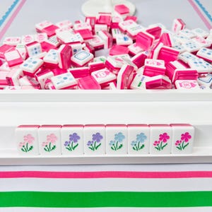 May include: A pink and white Mahjong set with a tray of tiles. The tiles are white with pink accents and symbols. The bottom row of tiles features floral designs in pink, purple, and blue.