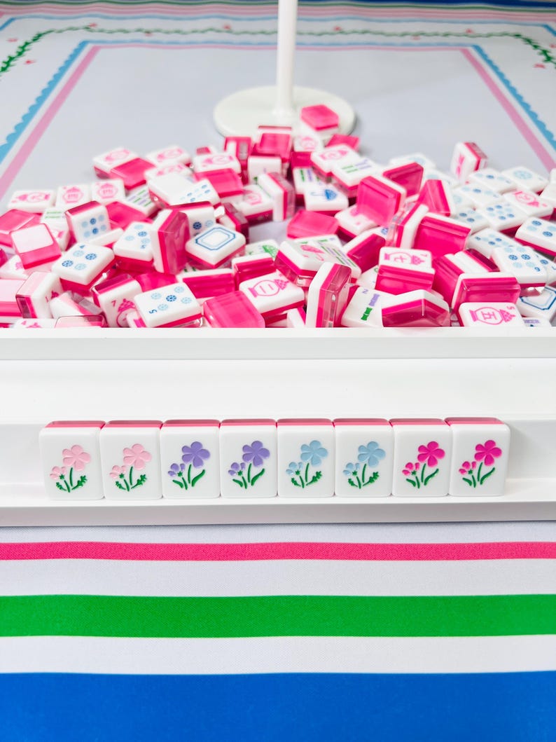 May include: A pink and white Mahjong set with a tray of tiles. The tiles are white with pink accents and symbols. The bottom row of tiles features floral designs in pink, purple, and blue.
