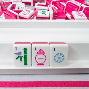May include: A collection of white and pink Mahjong tiles, with various symbols and numbers. Three tiles are in focus, displaying a bird, bamboo, and a vase. The tiles are arranged in a white tray, with a striped pink and green fabric in the background.