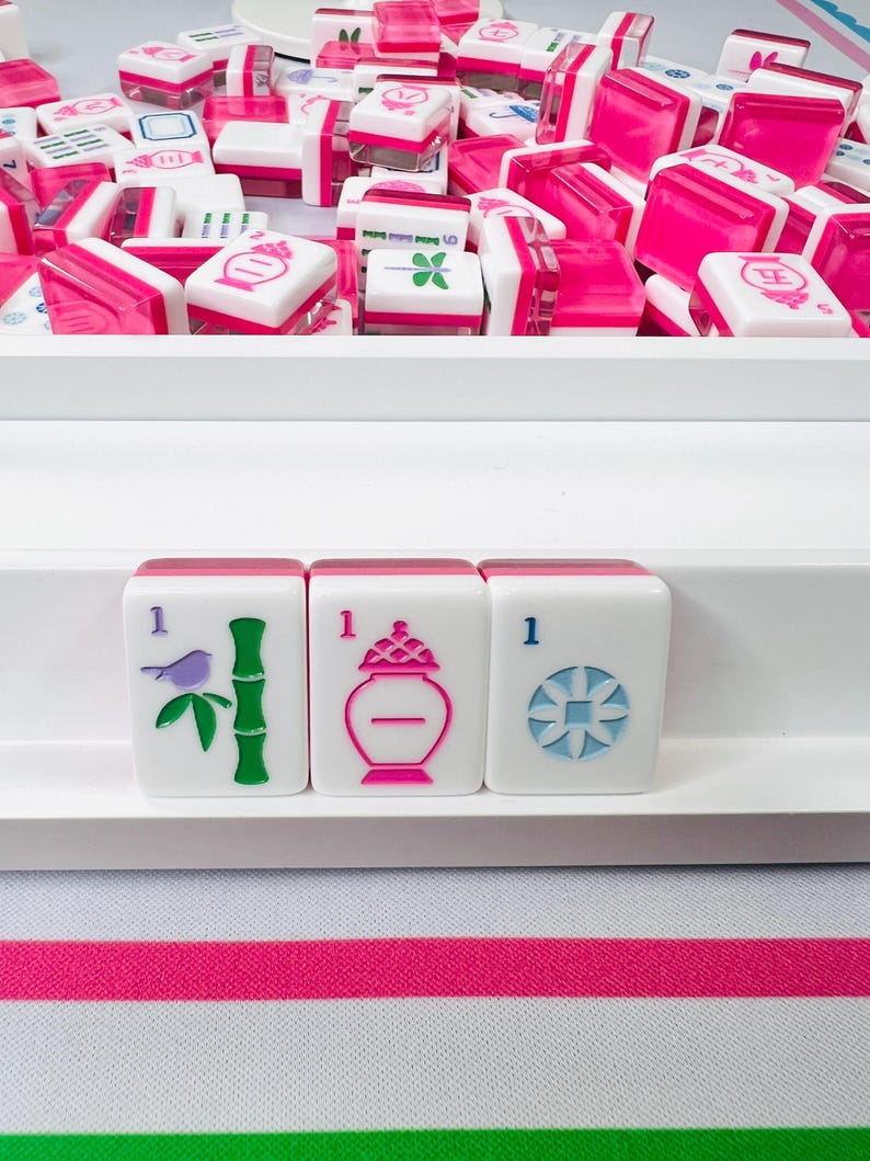 May include: A collection of white and pink Mahjong tiles, with various symbols and numbers. Three tiles are in focus, displaying a bird, bamboo, and a vase. The tiles are arranged in a white tray, with a striped pink and green fabric in the background.