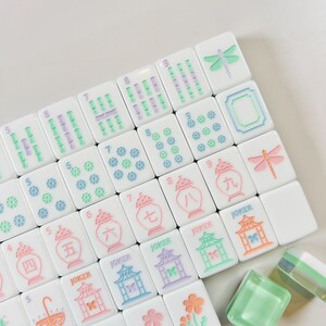 May include: White Mahjong tiles with colorful illustrations and numbers. The tiles feature images of flowers, dragonflies, and pagodas, with the word "JOKER" on some. The tiles are arranged on a white surface.