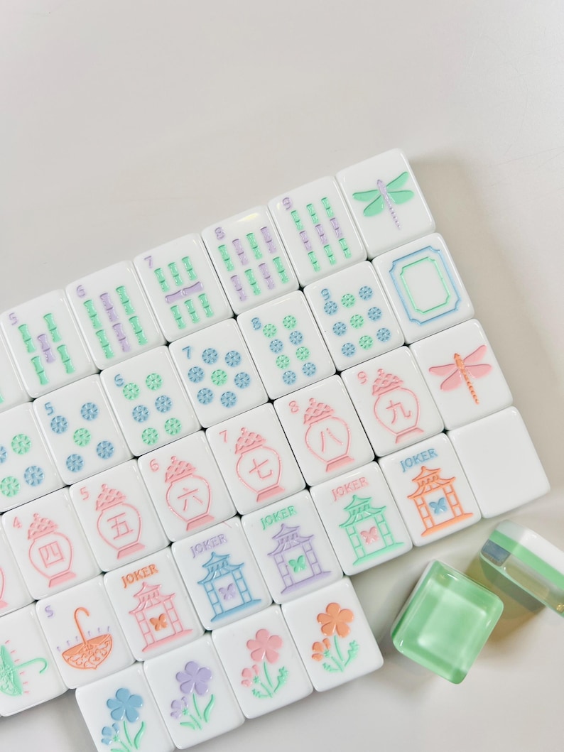 May include: White Mahjong tiles with colorful illustrations and numbers. The tiles feature images of flowers, dragonflies, and pagodas, with the word "JOKER" on some. The tiles are arranged on a white surface.