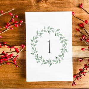 May include: A white card with a green leafy wreath and the number "1" in the center. The card is surrounded by red berry sprigs on a wood background.