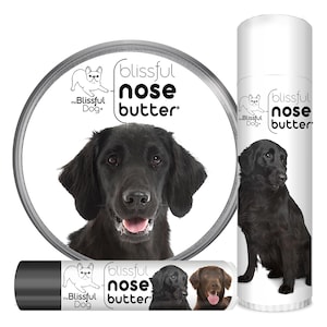May include: A black dog with a pink nose is featured on a round label with the text "blissful nose butter" and a small illustration of a dog. The label is attached to a tube of nose butter. The tube is black with white text that reads "blissful nose butter" and a small illustration of a dog. The tube is next to a larger tube of nose butter with the same design. The larger tube is white with black text.