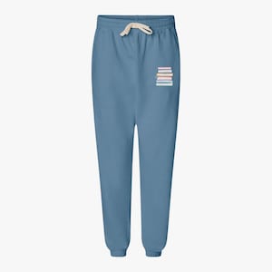 May include: Blue sweatpants with a drawstring waist and elastic cuffs. A small graphic of a stack of books in various colors is printed on the left leg. The pants are made of a soft material.
